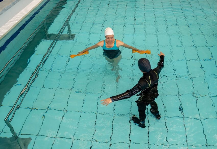 A person wearing a white swim cap extends their arms forward in a swimming pool while a therapist in black attire gently guides the movement, demonstrating a hydrotherapy balance or mobility exercise.