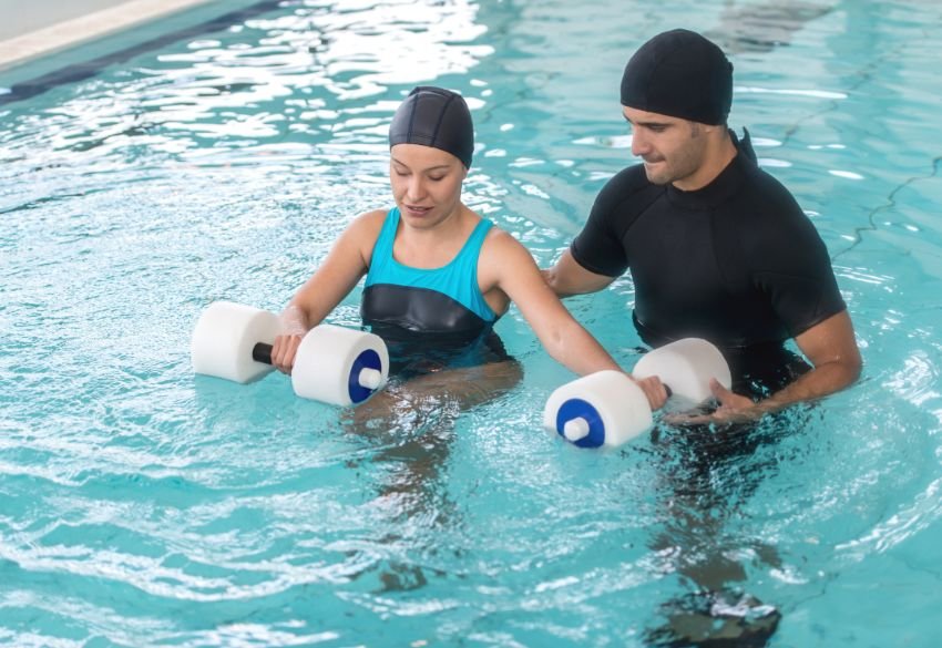 A woman in a blue swimsuit and black swim cap holds foam dumbbells in the water, performing resistance exercises with the assistance of a therapist in black attire.