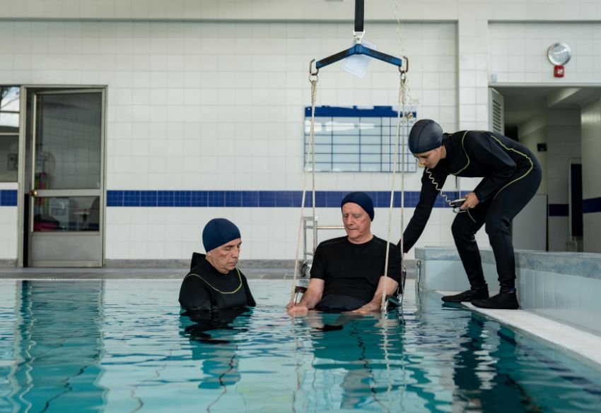 Three individuals in a pool participate in a hydrotherapy session; one person is seated in the water while two assistants support and prepare them for aquatic rehabilitation.
